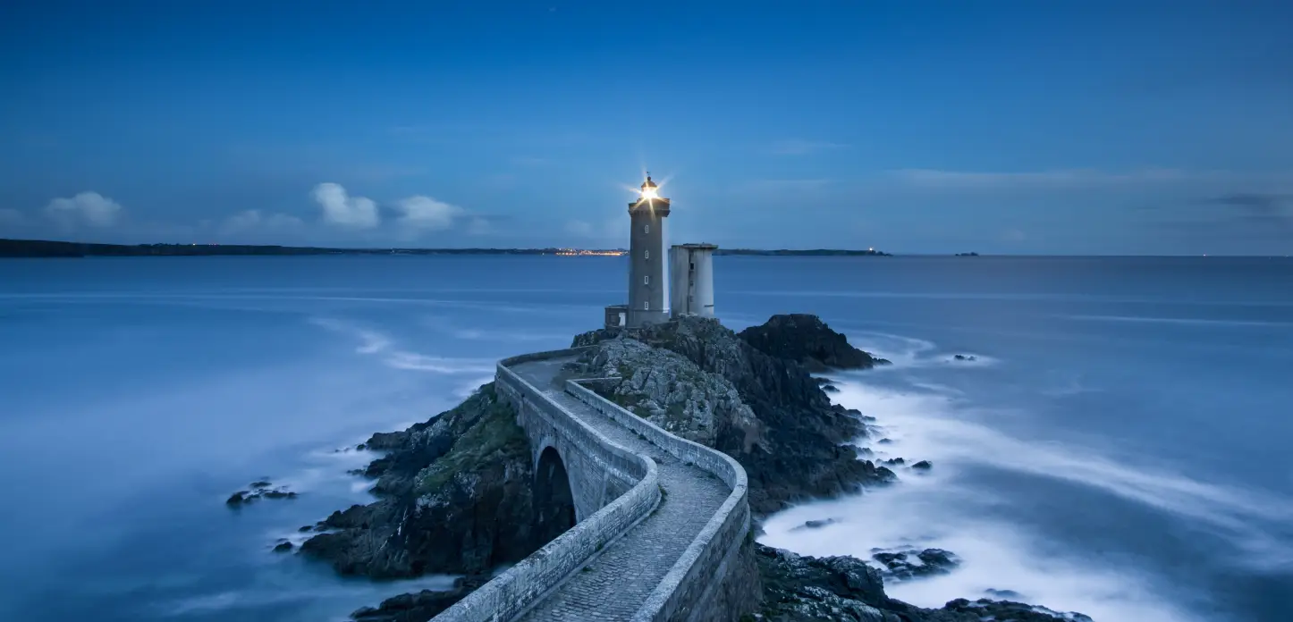 A lighthouse on a rocky islet at dusk, reached by a stone causeway