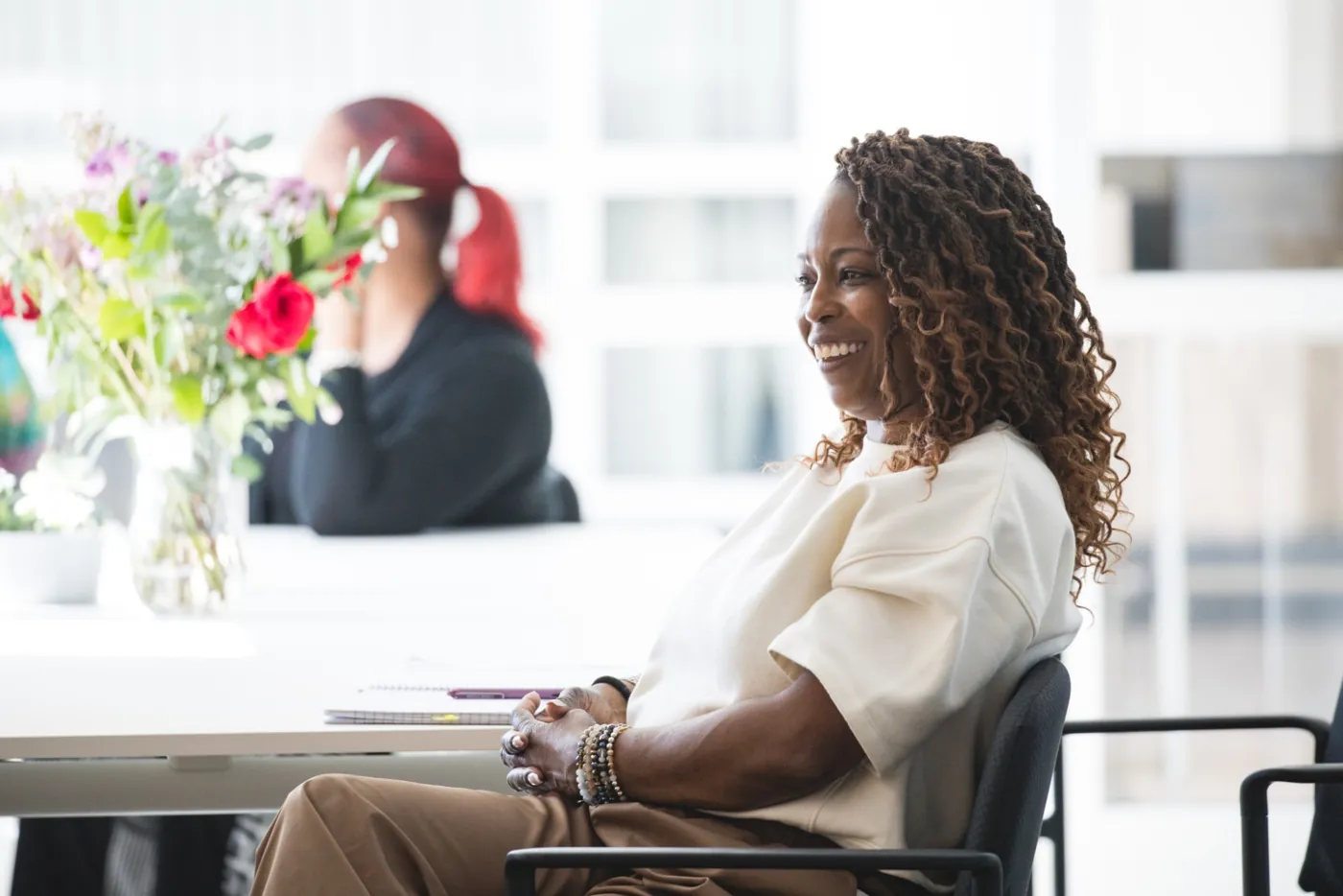 A smiling woman with long curly hair seated at a table in a bright classroom