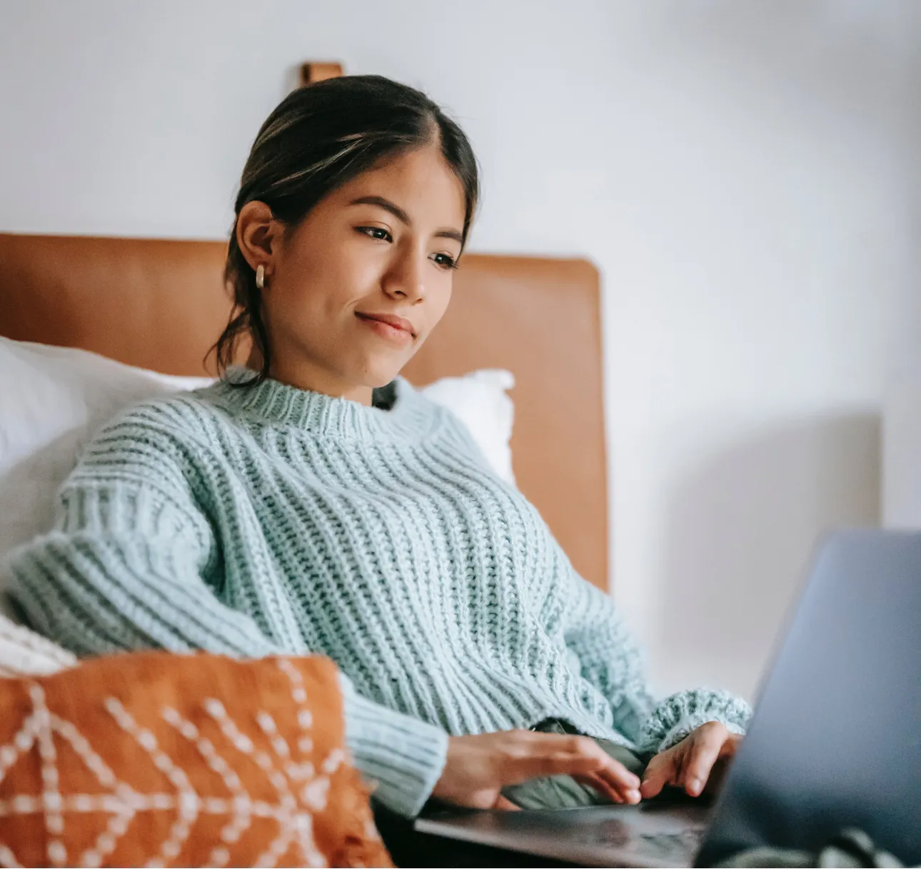 A woman sitting on her bed in a light blue sweater working on her laptop.