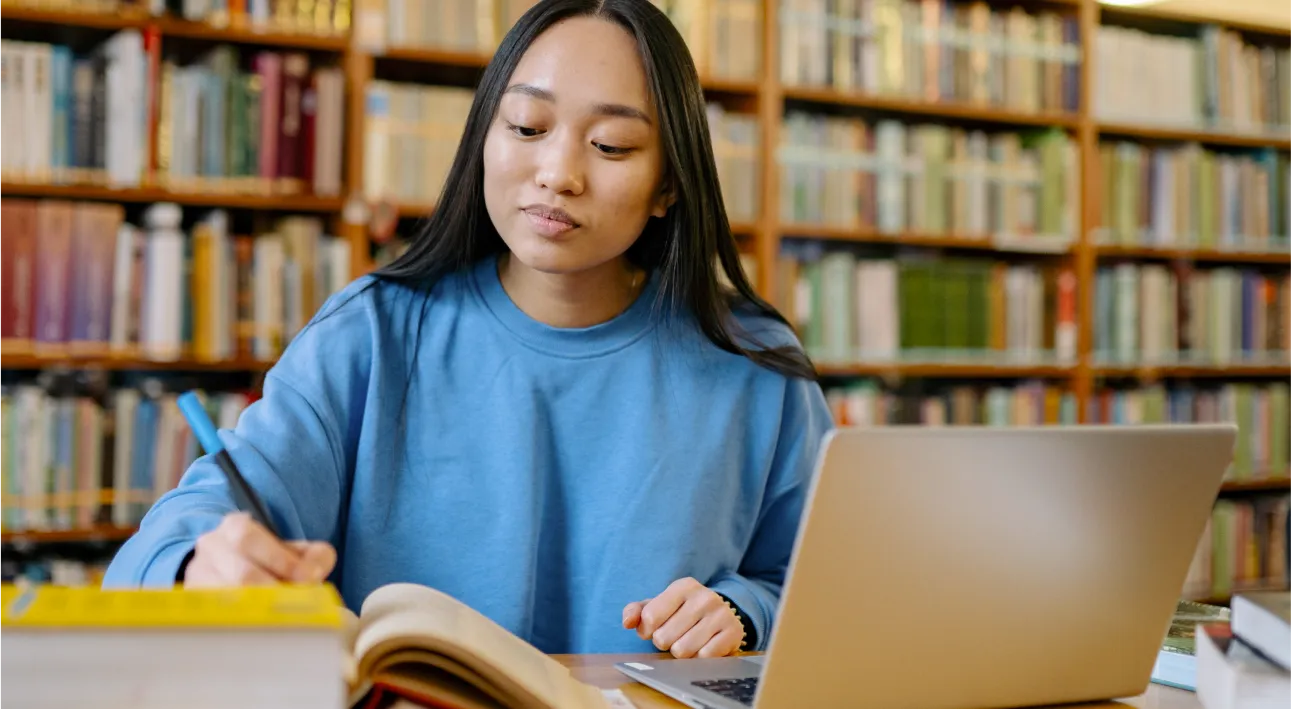 young woman sits behind laptop and book doin research in library