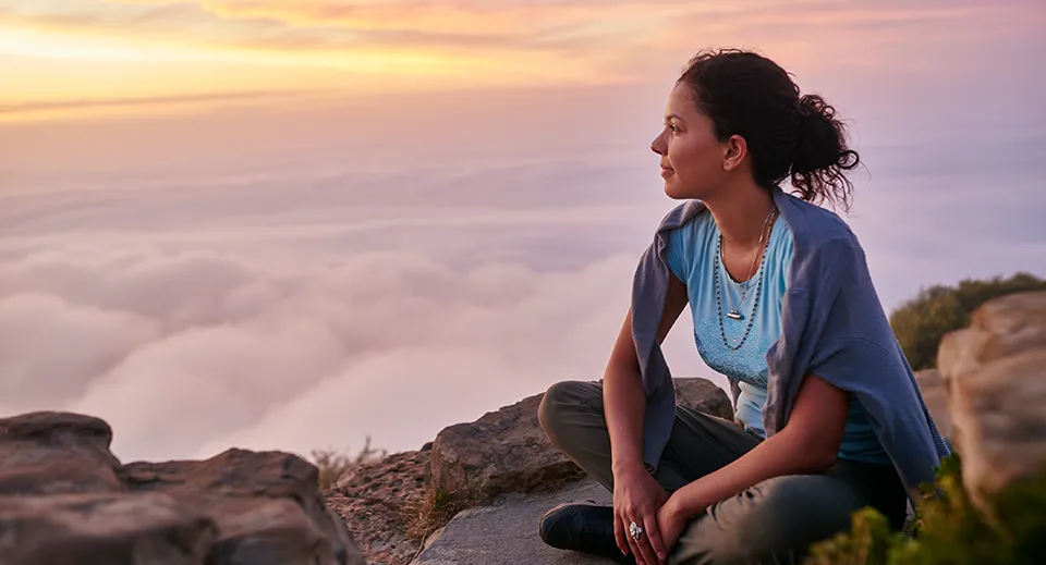Person sitting on a rocky overlook at sunrise above the clouds