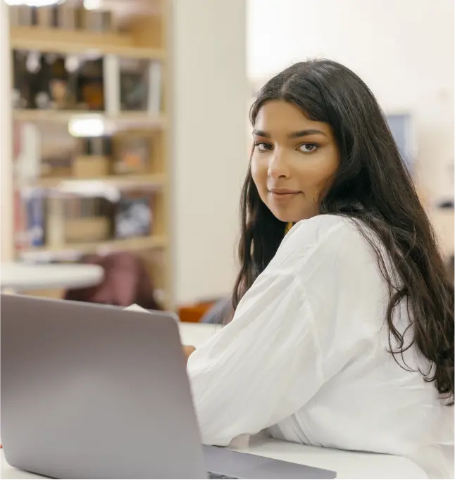 Woman looks over her shoulder in the library while sitting at desk with laptop