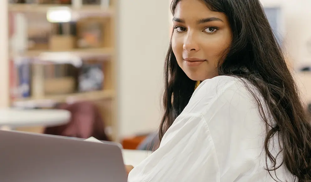 Woman looks over her shoulder in the library while sitting at desk with laptop