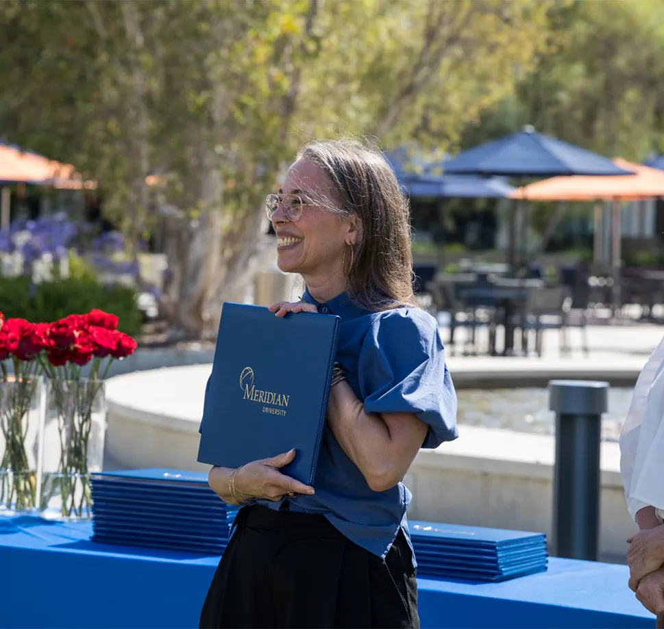 A smiling Meridian graduate holding a Meridian University diploma cover