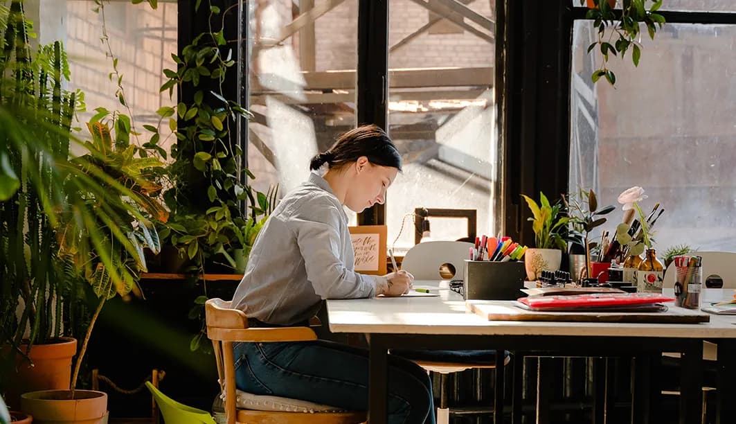 A person writing at a desk surrounded by plants