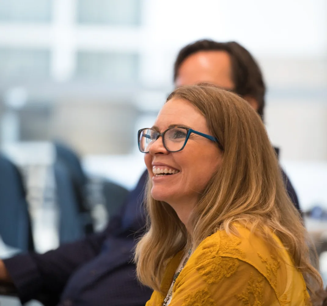 A student in glasses and a mustard-yellow top laughs during a class session, seated beside classmates