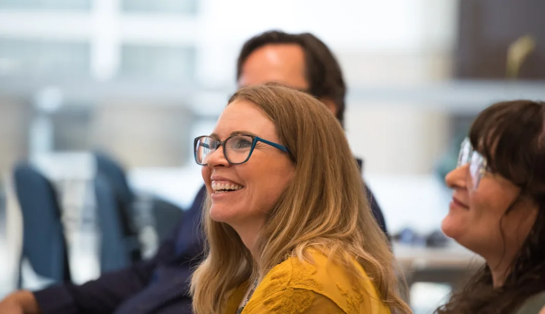 A student in glasses and a mustard-yellow top laughs during a class session, seated beside classmates