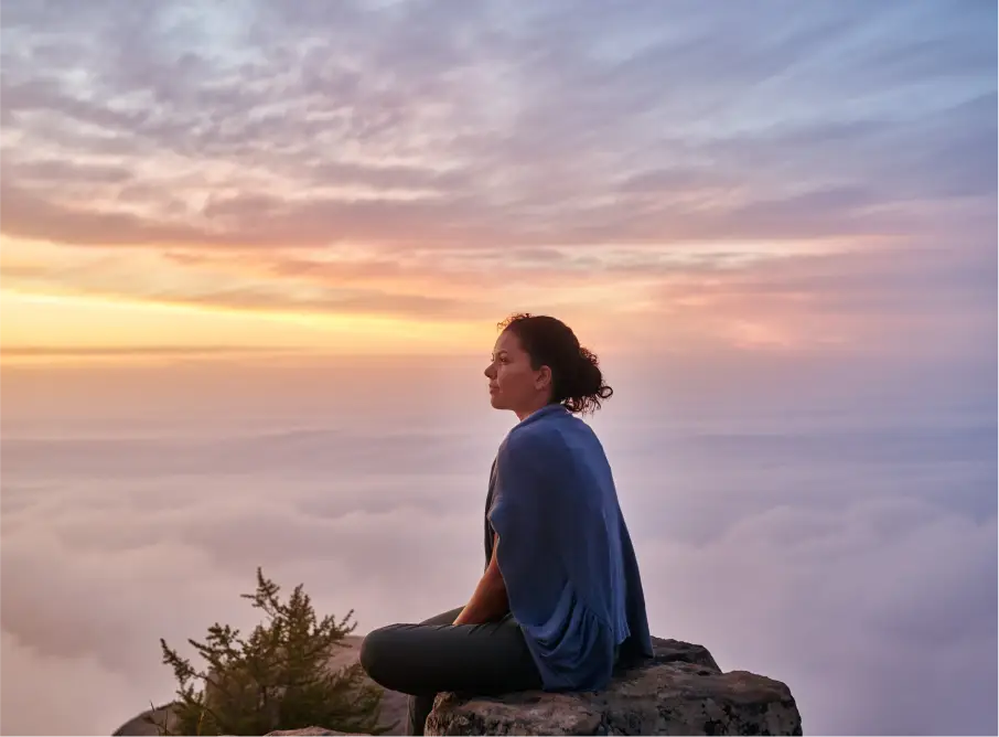 A woman sits cross-legged on a rocky outcrop above a sea of clouds, looking out at the sunrise