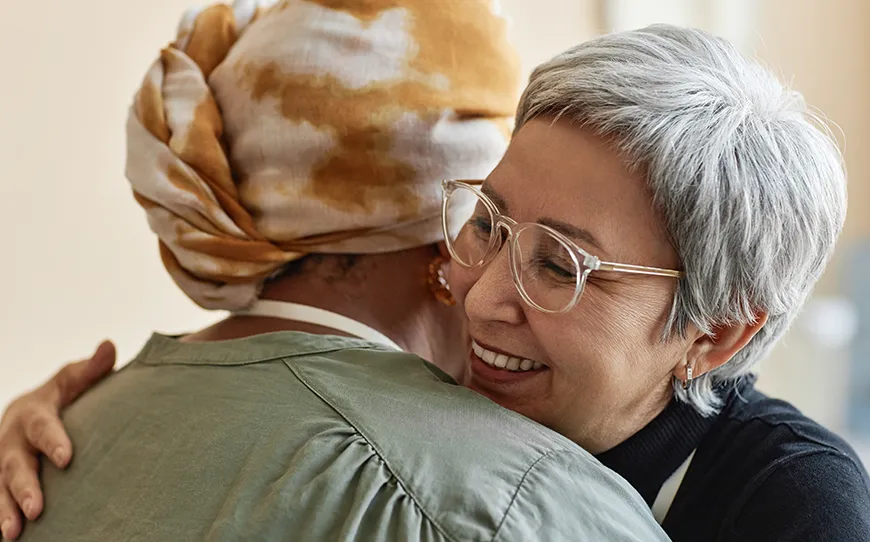 A warm hug between two women, one wearing glasses and smiling, the other in a patterned headscarf, capturing a moment of connection, trust, and support.