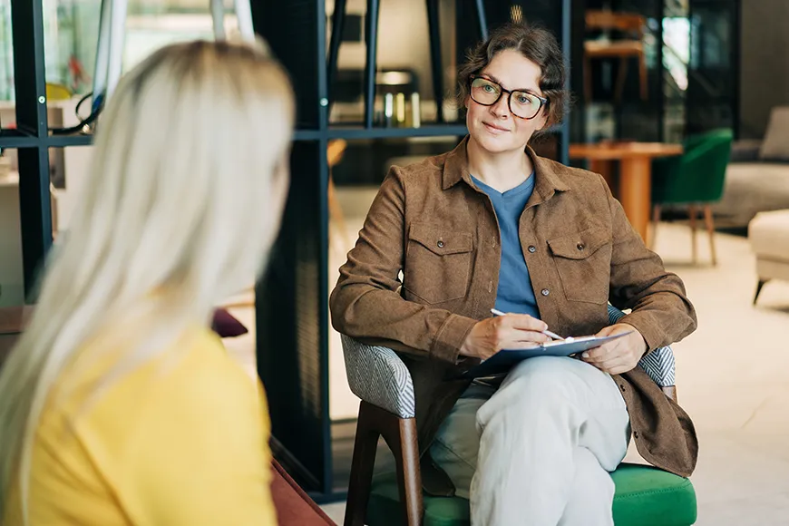 A female psychologist sits in a modern office holding a clipboard and listening attentively to a client during a therapy session.
