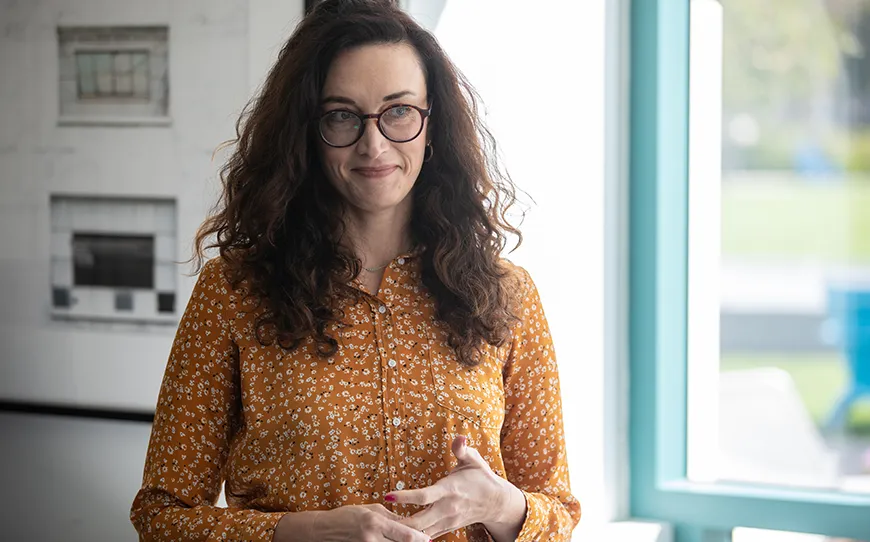 Woman with wavy dark hair and glasses, wearing a mustard yellow floral blouse, standing indoors near a bright window and smiling thoughtfully.