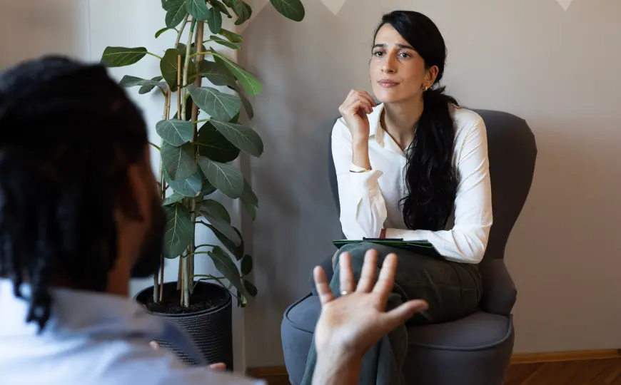 Female counselor in a white shirt holding a clipboard while listening to a male client during a counseling session in a softly lit office.