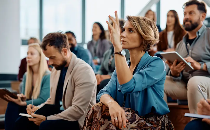 A professional raises her hand during a seminar while seated among other attendees taking notes.
