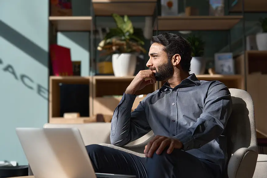 A young thoughtful savvy male professional executive looking away, relaxing, sitting on a chair in a modern office lobby with a laptop, thinking of new ideas, dreaming of success