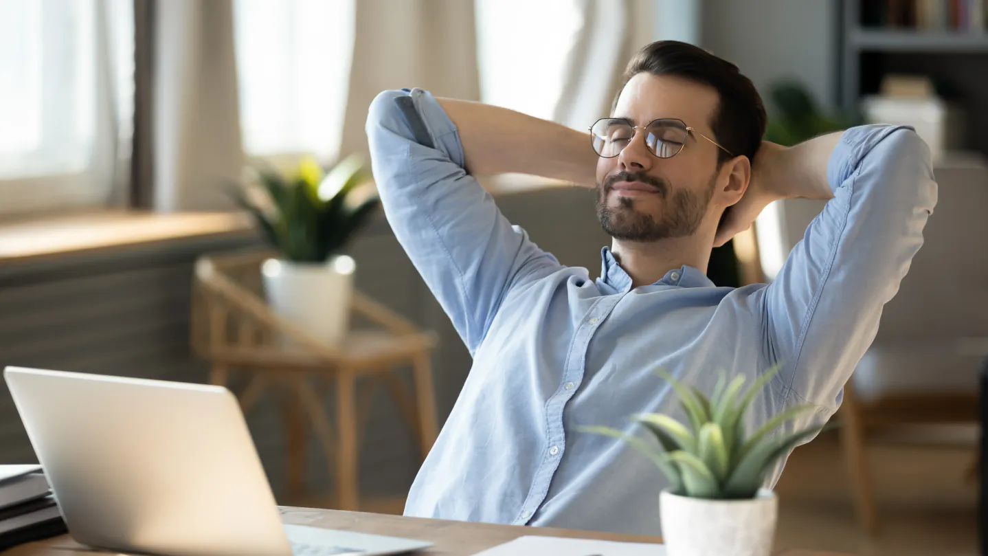 A man with glasses sits at a computer and leans back on his chair. He smiles and looks relaxed.