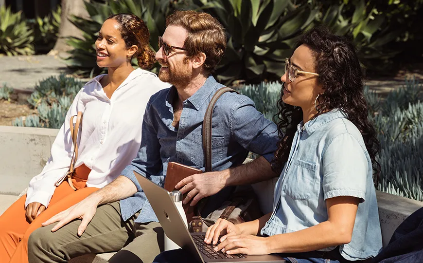 Three students sit together outdoors, one holding a laptop, engaged in conversation and collaboration on a sunny day.