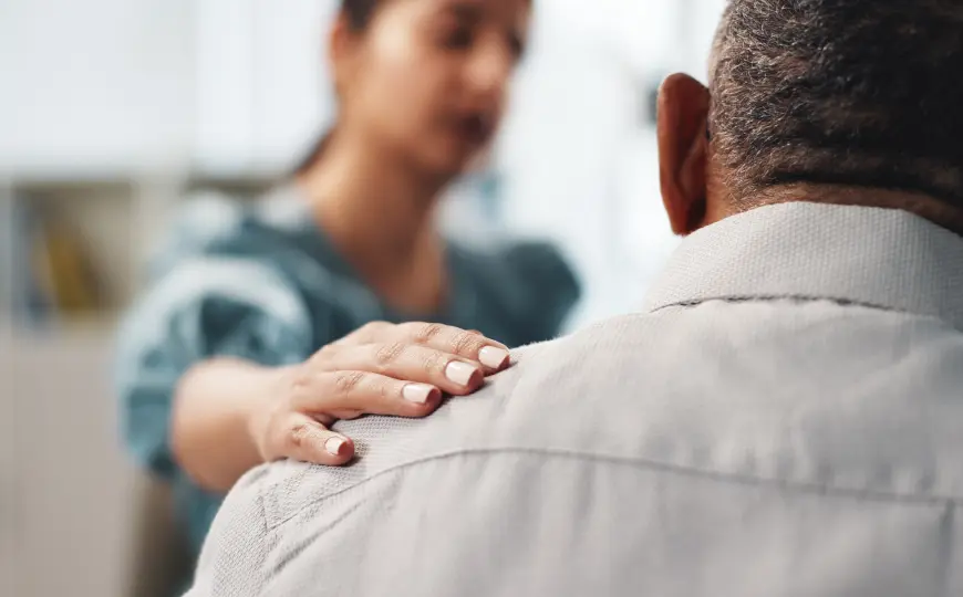 A therapist places a hand on a client's shoulder during a supportive therapy session focused on emotional presence and embodied care.