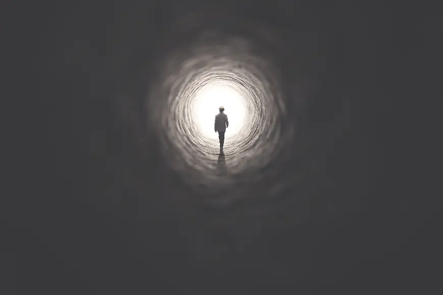 A silhouette of a person standing in the middle of a luminous field encased in a dark tunnel.