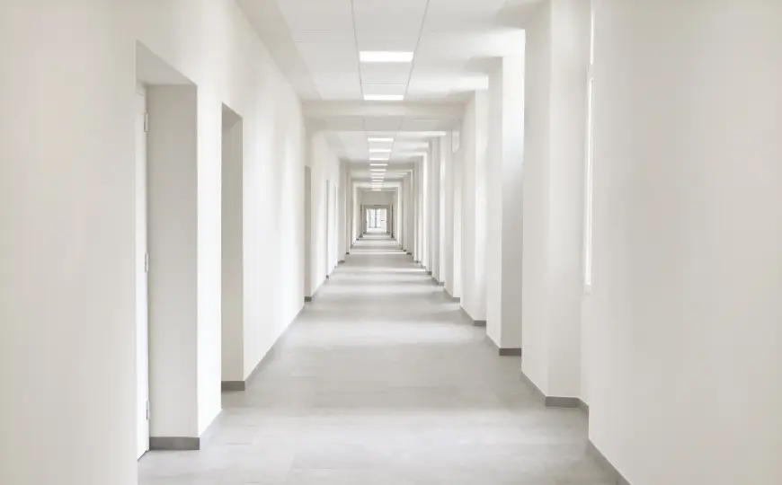 A long, empty white hallway with repeating doorways, bright ceiling lights, and a distant vanishing
