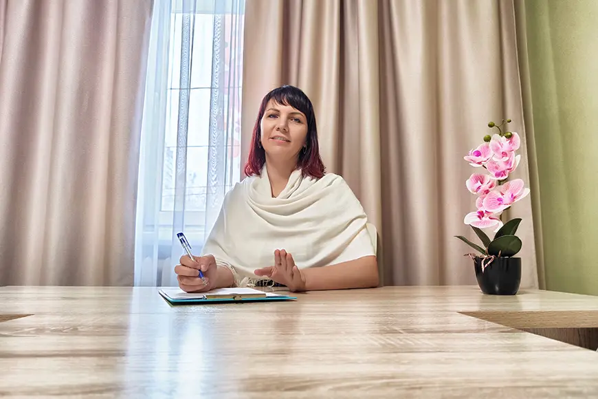 A counselor sits at a wooden desk with a notepad and pen, engaging in conversation during a session. Soft curtains and a potted orchid create a calm, welcoming atmosphere.