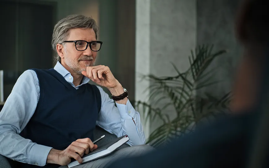 A thoughtful male therapist with glasses sits in a modern office, holding a notebook and pen while attentively listening during a therapy session.
