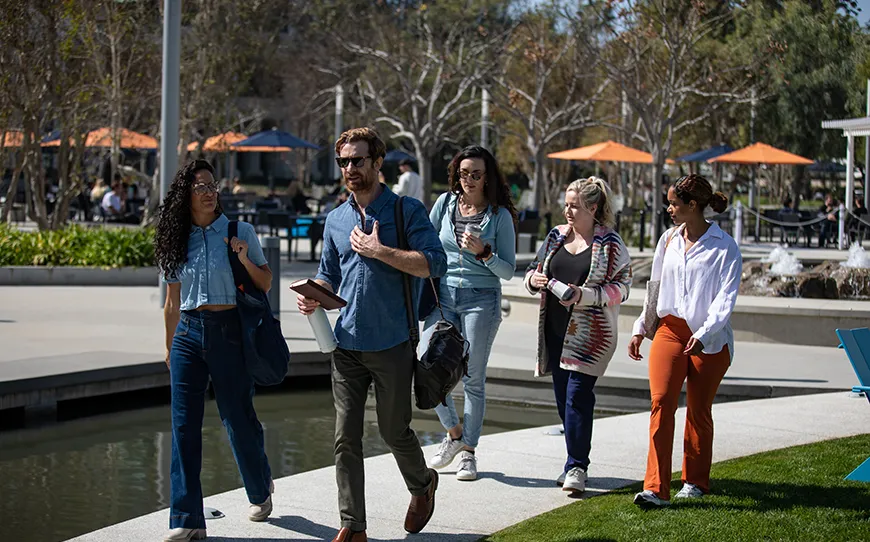 A group of five people walk together on a curved pathway beside a shallow water feature in a modern outdoor park.