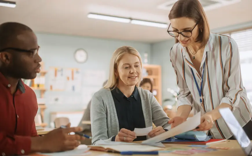 Three education professionals collaborate around a table reviewing documents together, one standing and sharing a paper with two seated colleagues in a bright classroom setting.