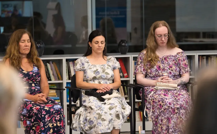 Three women seated with eyes closed and hands resting in their laps during a group meditation or contemplative practice, with bookshelves visible in the background.