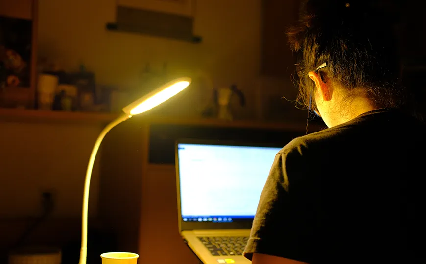 Student working late at night on a laptop under a desk lamp, focused on writing or studying.