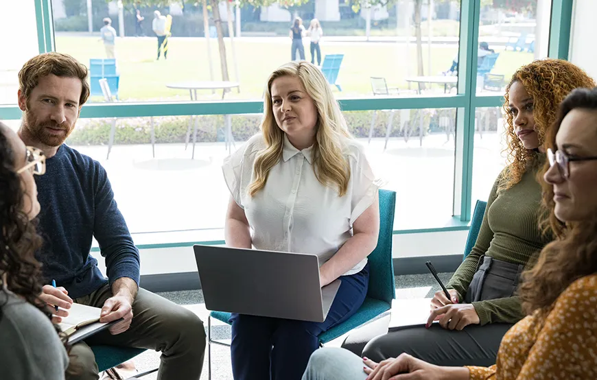 A group of adults in a professional setting sits in a discussion circle, with one woman holding a laptop and others taking notes, suggesting a collaborative or therapeutic meeting.