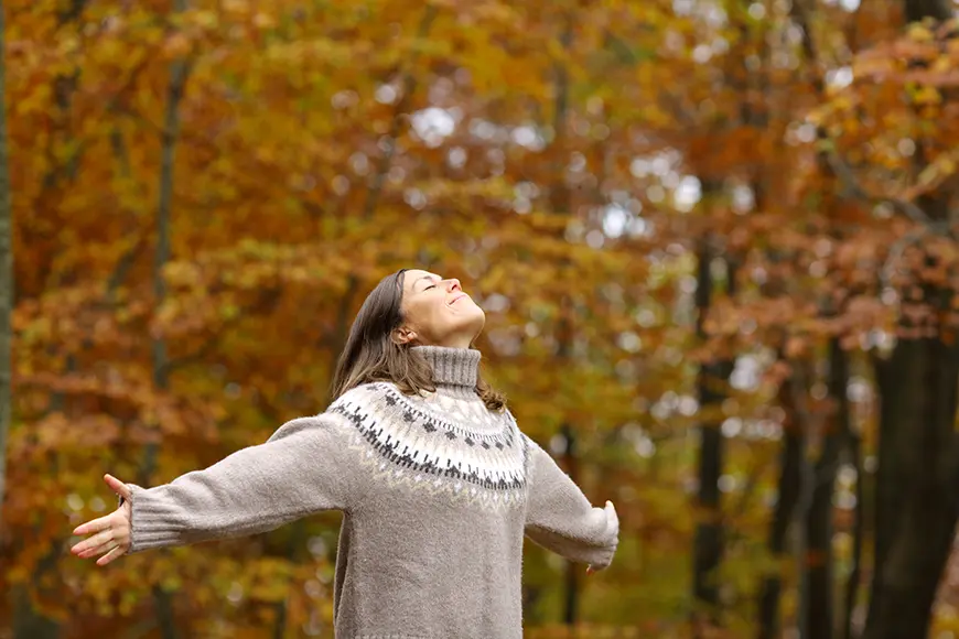 A woman who stands with her arms wide open in a forest area.