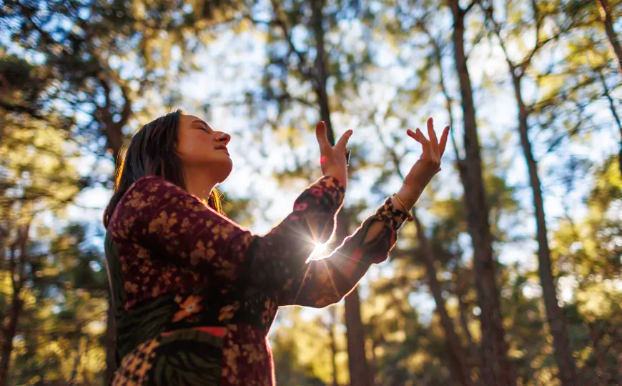 A person stands in a sunlit forest with eyes closed and hands raised in a reflective, body-aware posture.