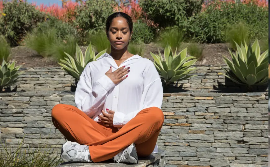 Person seated outdoors with a hand on the chest and abdomen during a calming breath practice.
