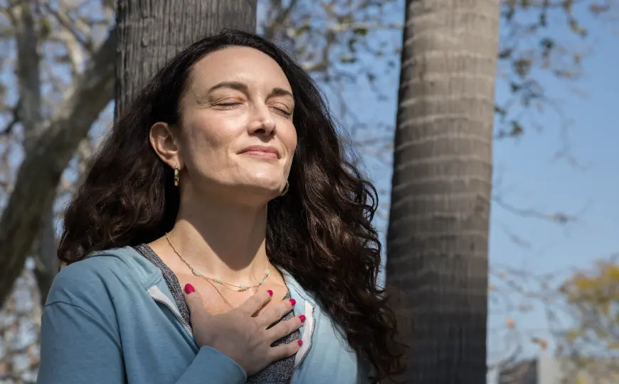Woman standing outdoors with eyes closed and hand on chest, practicing mindful breathing among trees.