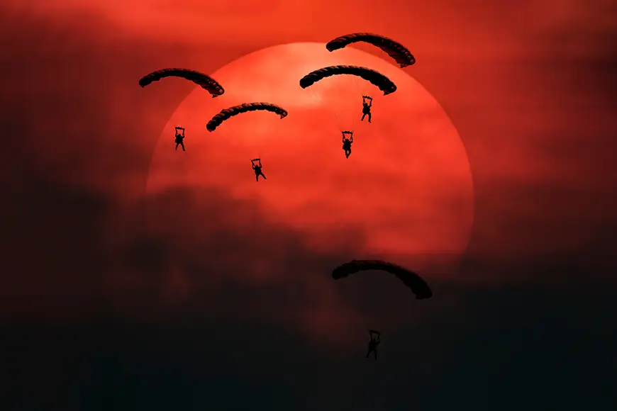 Five Special Operations parachutes silhouette in front of the sun with smoke and a battlefield mood