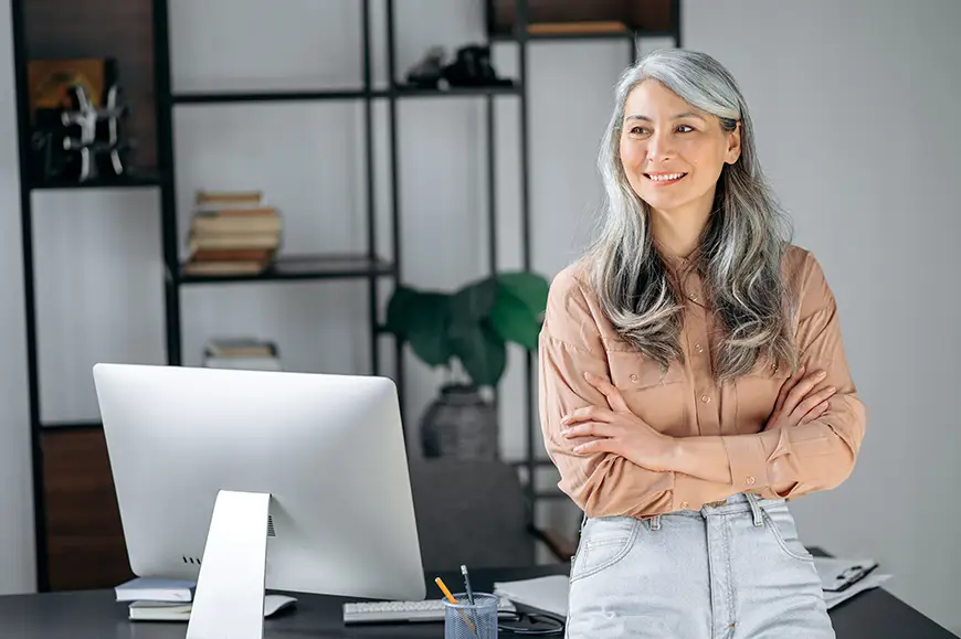 A business leader leans against a desk with arms crossed and a smile