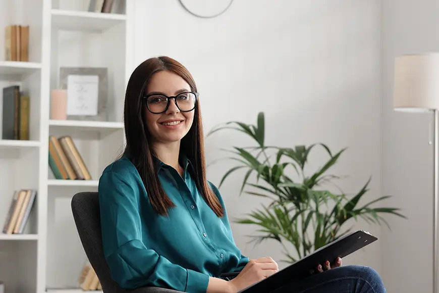 A smiling psychologist sitting in a chair while writing on a clipboard.