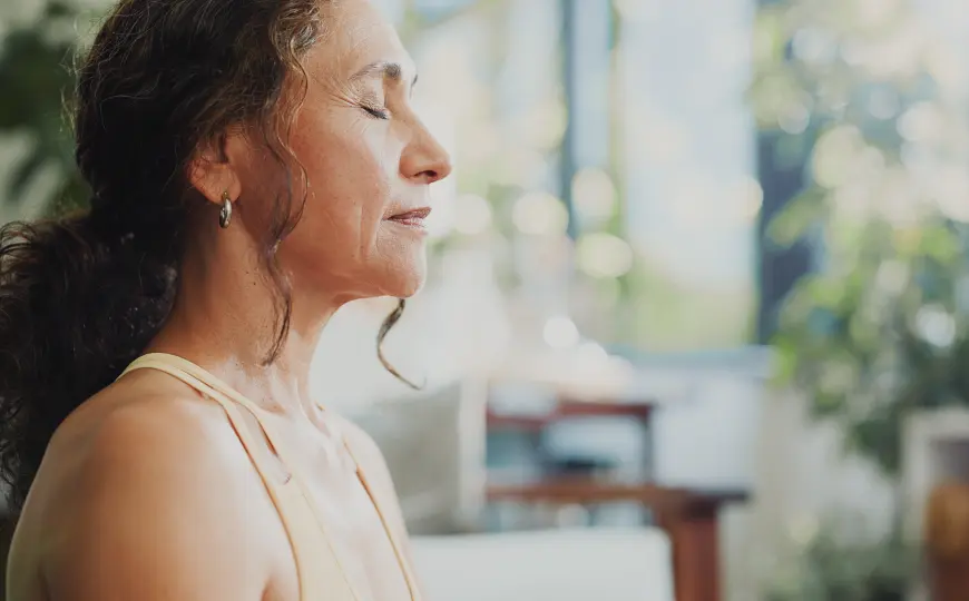Woman in profile with her eyes closed and a soft expression, seated in a bright room during a moment of quiet breath or meditation.