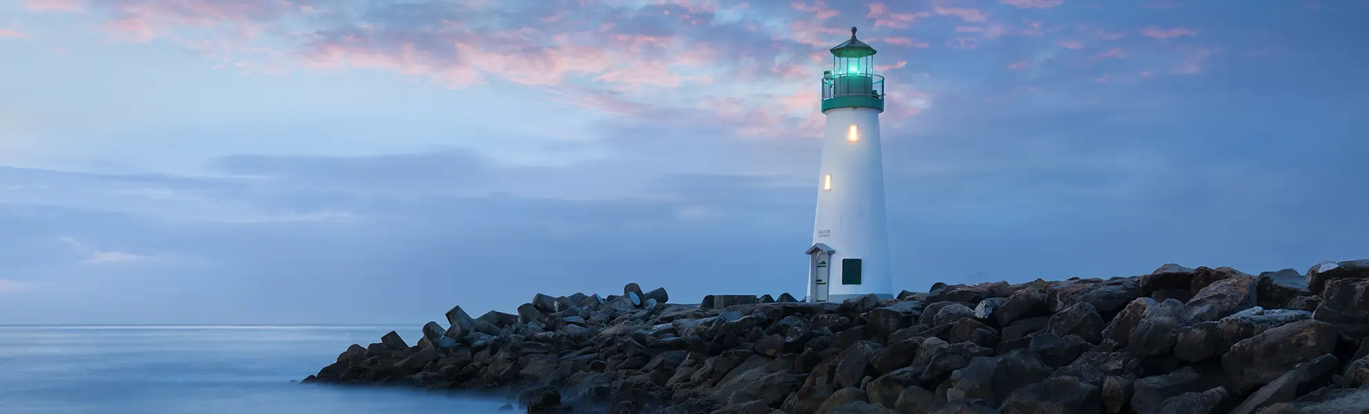A white lighthouse with a glowing green lantern on a rocky jetty at dusk